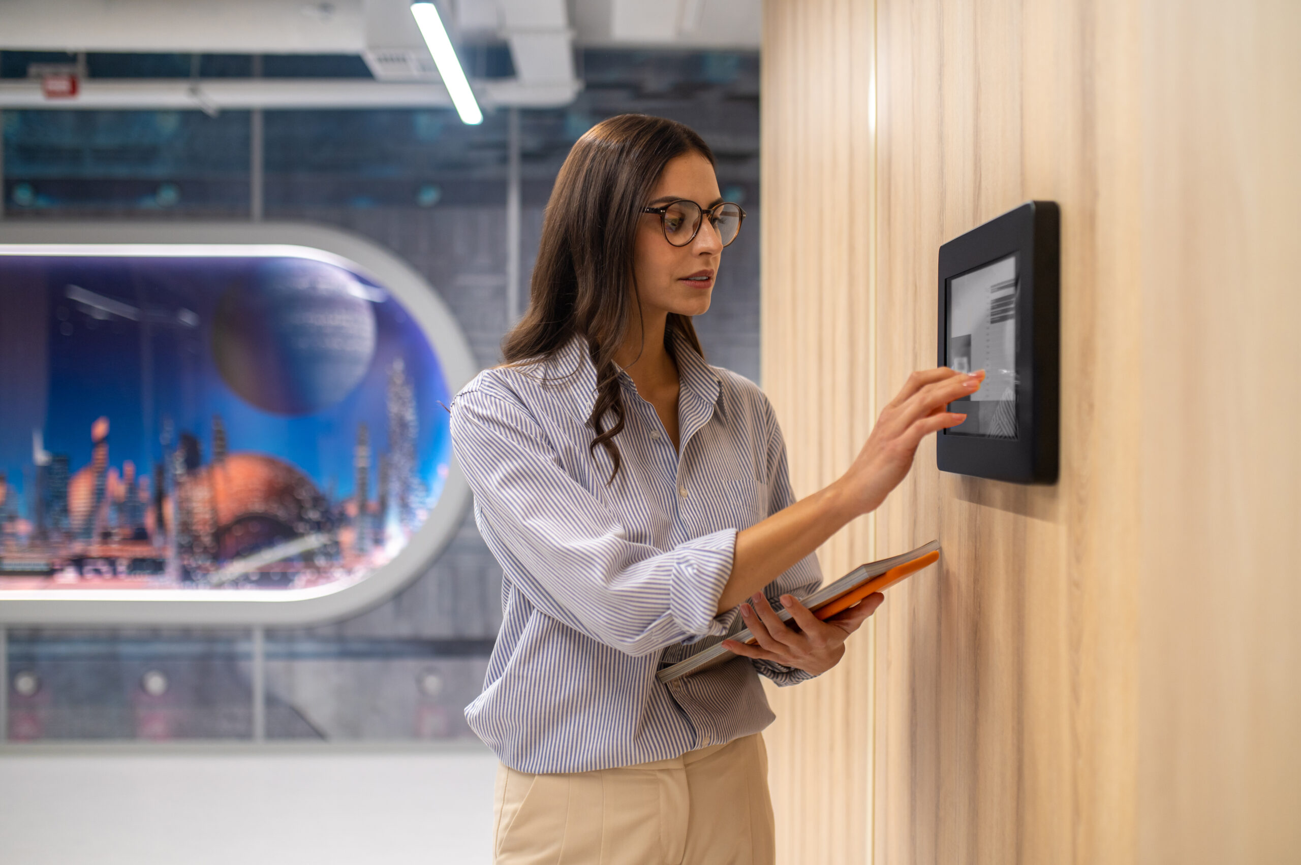 Woman touching control panel on wall in corridor Control panel. Focused elegant woman in glasses with folder touching with hand control panel on wall in illuminated corridor of school